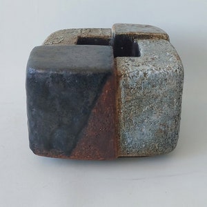 May include: A ceramic planter with a geometric design. The planter is made of three pieces that fit together to form a cube. The planter is glazed in shades of blue, gray, and brown.