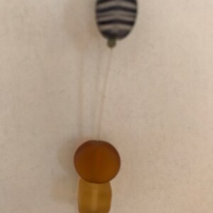 May include: Close-up of a necklace featuring a zebra-striped oval bead and a round amber bead atop a rectangular amber bead. The beads are strung on a thin, clear cord against a neutral background.