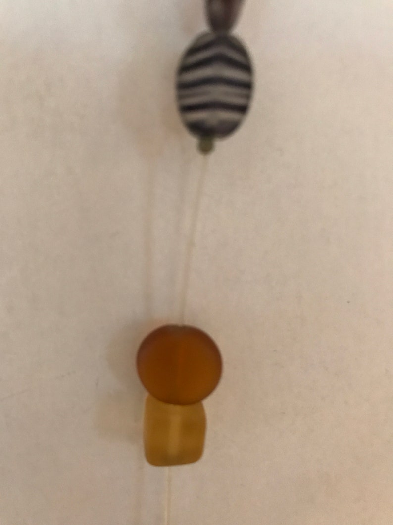 May include: Close-up of a necklace featuring a zebra-striped oval bead and a round amber bead atop a rectangular amber bead. The beads are strung on a thin, clear cord against a neutral background.