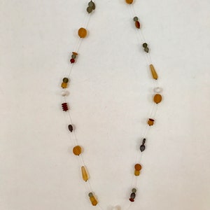 May include: A long necklace with various shaped beads in shades of amber, brown, and gray. The beads are strung on a clear, thin cord. The necklace features a mix of round, oval, and cylindrical beads.