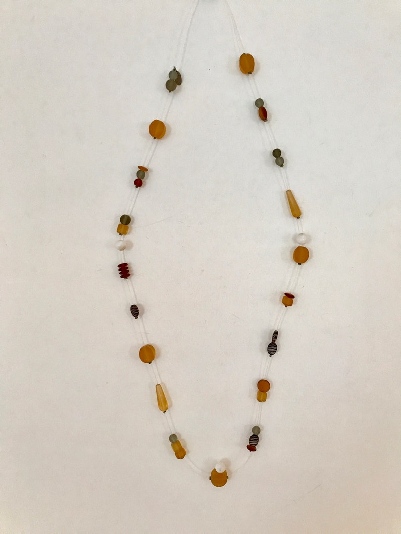 May include: A long necklace with various shaped beads in shades of amber, brown, and gray. The beads are strung on a clear, thin cord. The necklace features a mix of round, oval, and cylindrical beads.