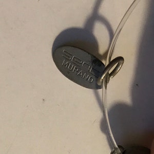 May include: Close-up of a silver-toned tag with the text "seni MURANO" attached to a clear, thin, flexible cord. A small, round, silver-toned bead is visible on the cord. The tag is oval-shaped and has a small loop.
