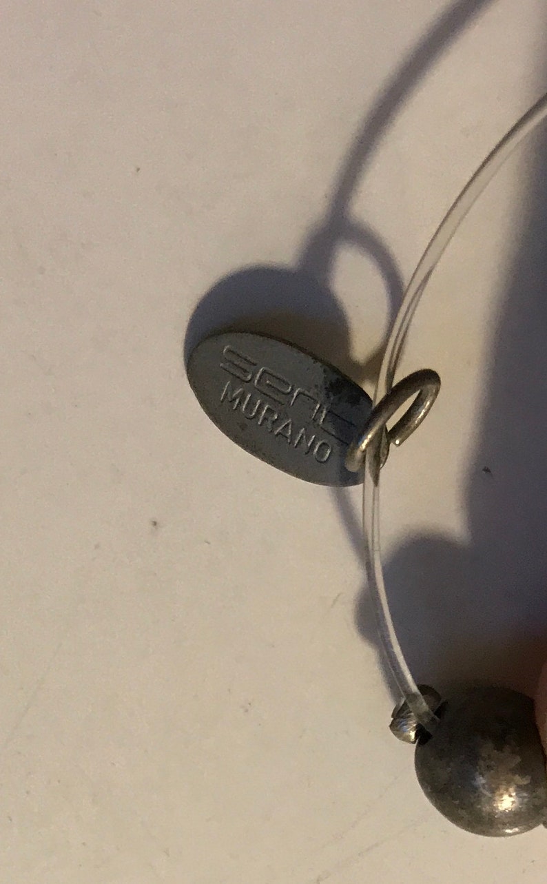 May include: Close-up of a silver-toned tag with the text "seni MURANO" attached to a clear, thin, flexible cord. A small, round, silver-toned bead is visible on the cord. The tag is oval-shaped and has a small loop.