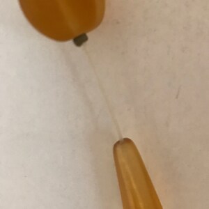 May include: A close-up of a single amber-colored bead necklace. The necklace features two amber-colored beads, one oval and one teardrop-shaped, connected by a thin, clear string. The beads have a smooth, polished surface.