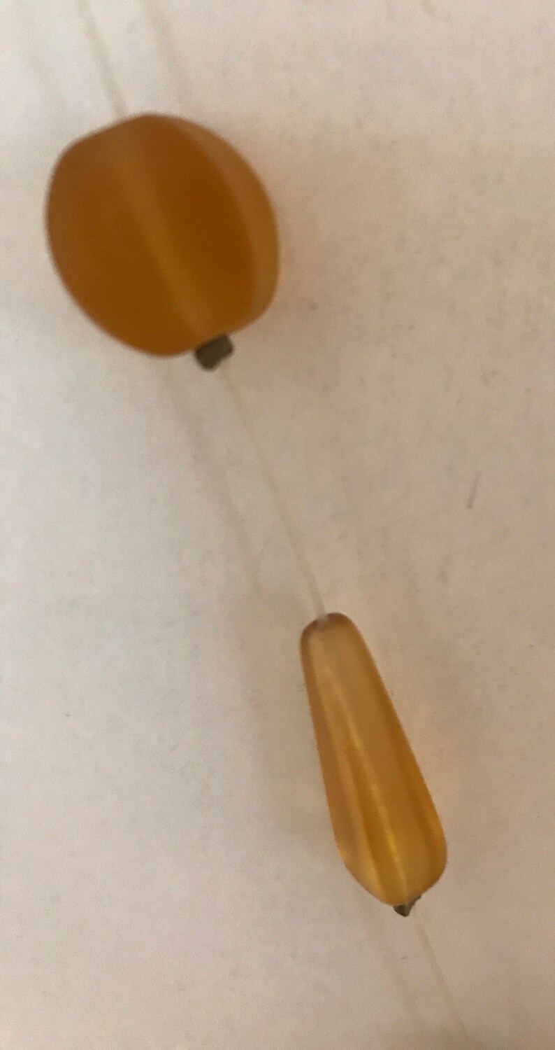 May include: A close-up of a single amber-colored bead necklace. The necklace features two amber-colored beads, one oval and one teardrop-shaped, connected by a thin, clear string. The beads have a smooth, polished surface.