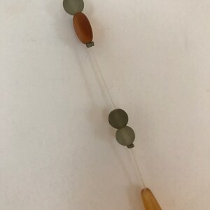 May include: A necklace with a clear string and various beads. The beads include a brown oval bead, two gray round beads, and a light orange teardrop-shaped bead. The beads are strung together in a linear fashion.