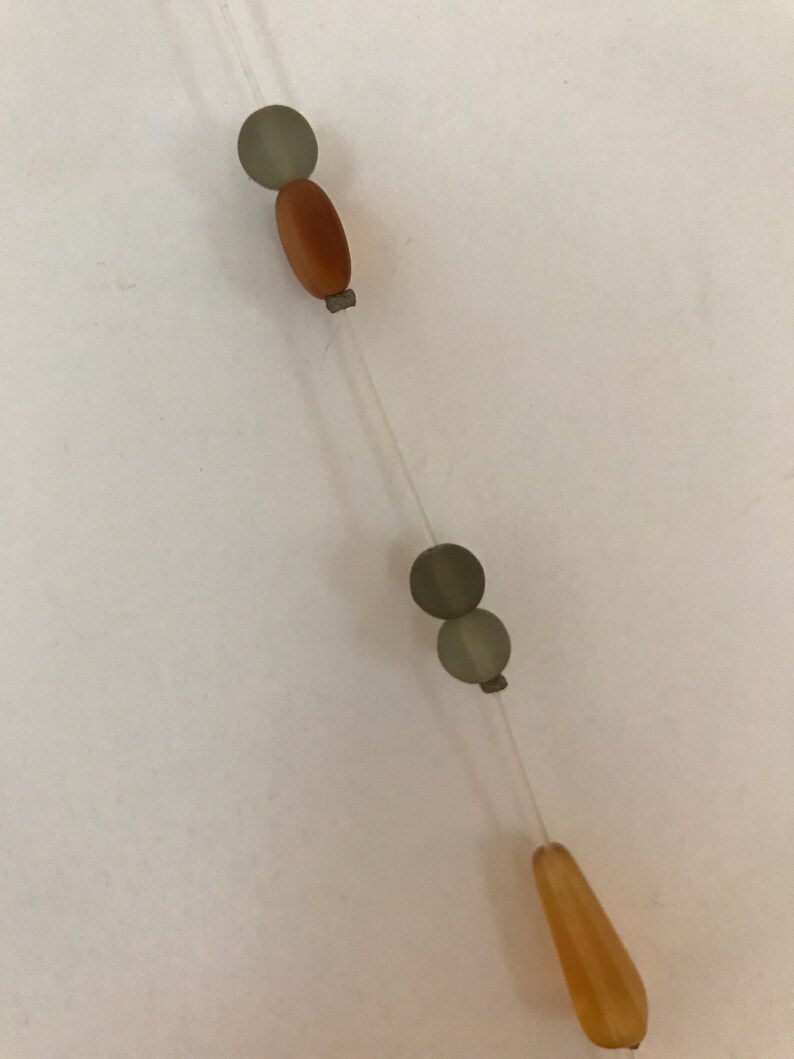 May include: A necklace with a clear string and various beads. The beads include a brown oval bead, two gray round beads, and a light orange teardrop-shaped bead. The beads are strung together in a linear fashion.