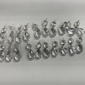 May include: A collection of clear, faceted glass chandelier crystals arranged in multiple rows. Each piece features a small circular bead connected to a larger teardrop-shaped pendant by a silver-toned metal jump ring, creating a dangling design.
