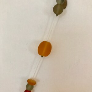 May include: A delicate necklace featuring a clear string with various beads. The beads are in shades of orange, green, and red. The necklace has a simple, minimalist design.