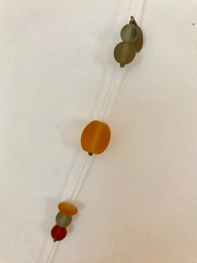 May include: A delicate necklace featuring a clear string with various beads. The beads are in shades of orange, green, and red. The necklace has a simple, minimalist design.