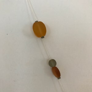 May include: A delicate necklace featuring a clear string with various beads. The beads include an oval amber-colored bead, a gray circular bead, and a brown oval bead. The necklace is simple and elegant.