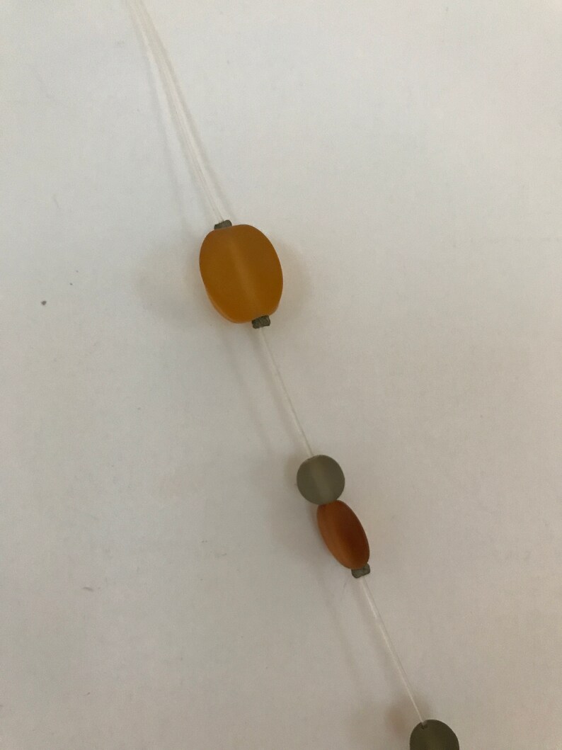 May include: A delicate necklace featuring a clear string with various beads. The beads include an oval amber-colored bead, a gray circular bead, and a brown oval bead. The necklace is simple and elegant.