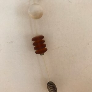 May include: A close-up of a necklace with various beads. The necklace features a mix of shapes and colors, including a rectangular amber bead, a white round bead, stacked brown beads, and a striped oval bead. The beads are strung on a thin, clear cord.