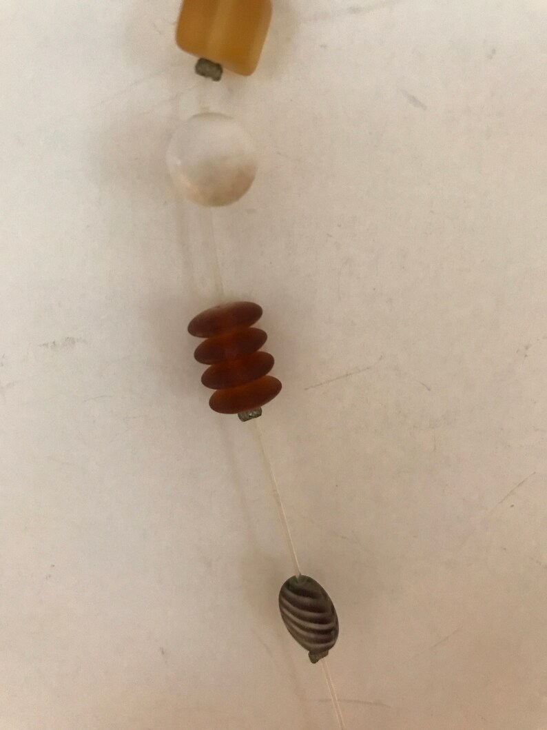 May include: A close-up of a necklace with various beads. The necklace features a mix of shapes and colors, including a rectangular amber bead, a white round bead, stacked brown beads, and a striped oval bead. The beads are strung on a thin, clear cord.