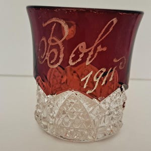 May include: A clear glass tumbler with a cut glass pattern. The top of the tumbler is a deep red color with the inscription "Bob 1911" in white.