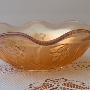 May include: An iridescent orange glass bowl with a ruffled rim. The bowl has a floral pattern embossed on the sides. The bowl is sitting on a white lace tablecloth.