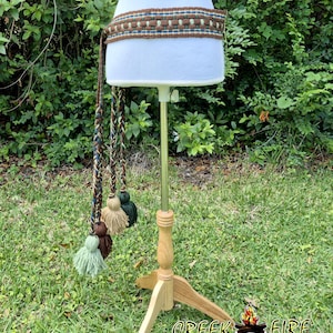 May include: A woven belt with brown, teal, and cream colors, displayed on a mannequin. The belt has a long braided tassel with multiple tassels in shades of green, brown, and cream. The logo "Creek Fire" is visible at the bottom.