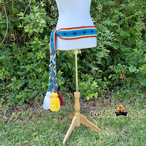 May include: A woven belt with a turquoise, red, and yellow pattern, displayed on a mannequin. The belt has a long, braided tassel in blue, yellow, and red, with white, yellow, and red tassels at the end. The belt is displayed on a wooden stand.