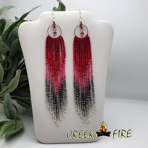 May include: A pair of long, beaded earrings with a gradient of red, pink, and gray beads. The earrings are hanging from silver hooks and have a small silver ring at the top.