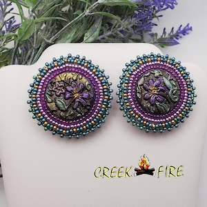 May include: A pair of round, teal, purple, and gold beaded earrings with a floral design in the center. The earrings are approximately 2 inches in diameter.