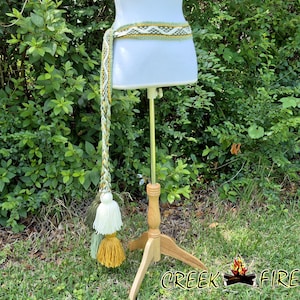 May include: A woven belt with a long tassel, displayed on a dress form. The belt features a geometric pattern in shades of green and white. The tassel includes white, green, and yellow tassels. The dress form is on a wooden stand, set against a backdrop of green foliage.