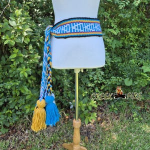 May include: A woven belt with a colorful geometric pattern in blue, yellow, and red. The belt has a long, braided tassel with yellow and blue tassels. The text "Creek Fire" is visible in the image.