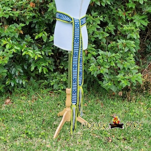 May include: A woven belt with a blue and white geometric pattern, accented with neon green and black fringe. The belt is displayed on a mannequin, showcasing its design and texture. The belt is a fashion accessory.