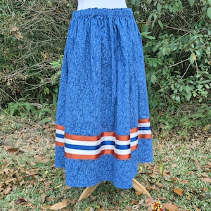 May include: A blue skirt with a patterned design and an elastic waistband. The skirt features horizontal bands of orange, white, and blue ribbon near the hem. The skirt is displayed on a wooden stand outdoors.