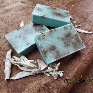 May include: Three bars of handmade soap with a light blue and brown marbled appearance. The soap is surrounded by dried sage leaves.