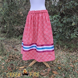 May include: A pink skirt with a floral pattern and an elastic waistband. The skirt features three horizontal bands of ribbon in white, teal, and burgundy. The skirt is displayed on a mannequin.