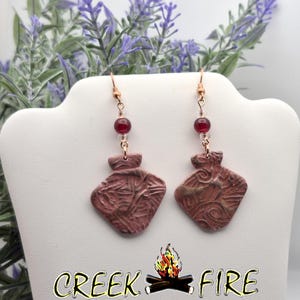May include: A pair of handcrafted earrings with a rustic, earthy aesthetic. Each earring features a textured, reddish-brown pendant with a unique design, suspended from a copper-colored hook. A small, red bead accents the top. The text "CREEK FIRE" is at the bottom.