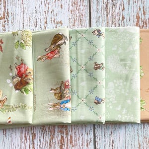 May include: Five folded fabric pieces with various patterns. The fabrics feature illustrations of rabbits, floral designs, and geometric patterns in shades of green and tan. The fabrics are arranged on a light blue wooden surface.