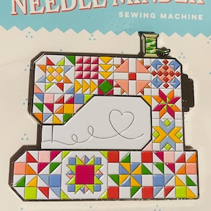 May include: A colorful "Quilty Needle Minder" in the shape of a sewing machine. The needle minder features a patchwork design with various colors and patterns. The text "Never lose another needle!" is at the bottom.