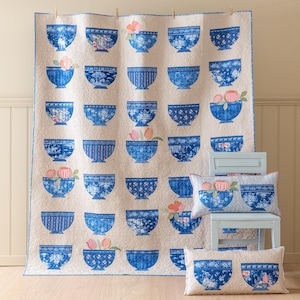 May include: A large quilt and two pillows feature a repeating pattern of blue bowls with floral designs. The quilt is cream-colored with blue bowls, some holding pink flowers. The pillows echo the bowl design.
