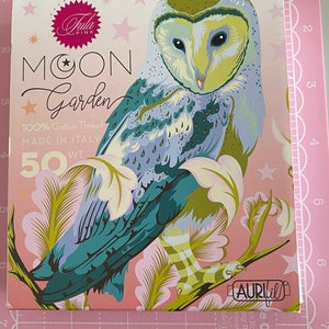 May include: A pink and green owl with a white face and yellow eyes is printed on a pink background with white stars. The text "Jula Pink Moon Garden 100% Cotton Thread Made in Italy 50 WT" is printed on the card. The text "Aurifil" is printed in the bottom right corner.