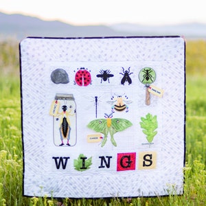 May include: A white quilt with a black border and a white background. The quilt features a variety of embroidered insects, including a ladybug, a fly, a bee, a moth, and a cricket. The quilt also features the word "WINGS" in large, colorful letters.