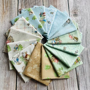 May include: A collection of folded fabric squares in various pastel colors, featuring illustrations of rabbits and text. The fabrics include light blue, green, and beige, with patterns of bunnies, foliage, and text. The arrangement is circular.