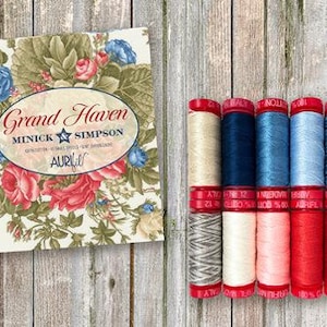 May include: A set of six spools of thread in various shades of blue, red, and white. The spools are labeled "Aurifil" and are arranged in a row on a wooden surface. The text "Grand Haven Minick & Simpson" is visible on a card behind the spools.