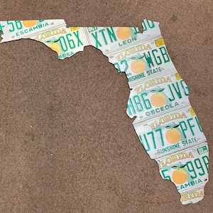 May include: A Florida state outline made of license plates. The plates are arranged to form the shape of the state. Each plate has a different county name and a different license plate number. The plates are all white with green lettering and a picture of an orange.