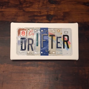 May include: A white framed sign with the word "DRIFTER" spelled out using colorful license plates. The letters are in various shades of blue, green, red, and black.