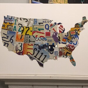 May include: A white canvas with a cutout of the United States of America. The cutout is filled with vintage license plates from different states. The license plates are arranged to form the shape of the United States.