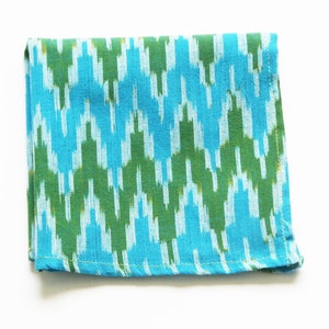 Op de afbeelding: Een blauwe en groene gepatroneerde stoffen servet met een witte achtergrond. Het servet heeft een geometrisch ontwerp en is dubbelgevouwen.