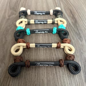 Puede incluir: Cuatro piezas de aparejo para caballos de cuerda, cada una con una combinación de colores diferente. La superior es negra y beige, la segunda es beige y verde oliva, la tercera es negra y turquesa, y la inferior es negra y beige. Cada una tiene el texto "SaddleTitty Fun".