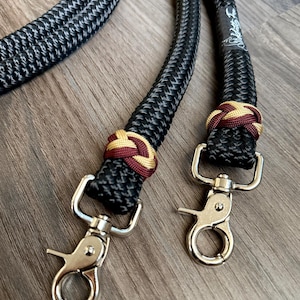 May include: Close-up of a black and gray rope leash with silver metal clasps. The leash features decorative knots in burgundy and gold. The background is a wooden surface.
