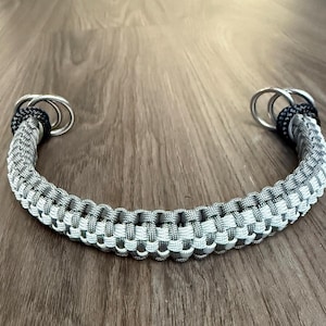 May include: A gray and white woven paracord handle with silver metal rings on each end. The handle is curved and appears sturdy, suitable for a bag or other accessory. The ends are secured with black cord.