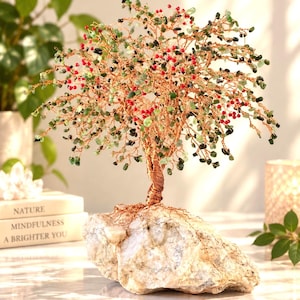May include: A decorative wire tree sculpture featuring twisted copper-colored wire branches adorned with small green stone chips and bright red beads. The tree is anchored to a large, irregular, light-colored natural stone base.