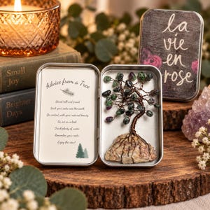 May include: A metal tin containing a wire-wrapped tree sculpture with green and purple stone leaves on a rock base. The opposite side features a card with the text Advice from a Tree and a list of inspirational phrases. The lid reads la vie en rose.