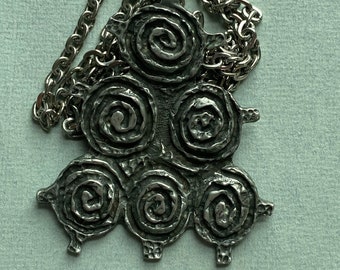 Puede incluir: Un collar de plata con un colgante con un diseño de seis círculos en espiral. El colgante está unido a una cadena de plata.