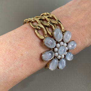 May include: A gold chain bracelet with a large white flower charm. The flower charm has a swirl pattern and is surrounded by smaller white pearls.