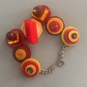 Puede incluir: Un brazalete de botones rojo y amarillo con cierre de cadena plateada. Los botones están superpuestos y tienen un aspecto vintage.